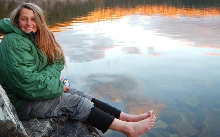 a person sits on a rock barefoot, with their feet dangling above still water that is reflecting the sky